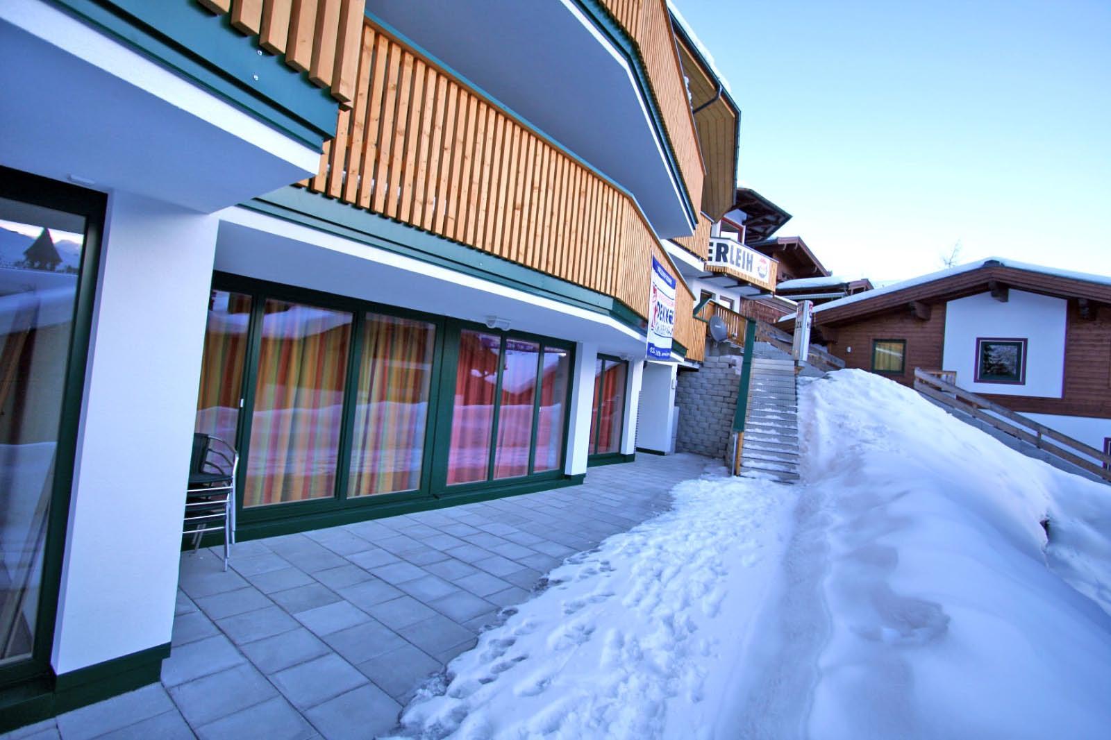 A modern building with wooden cladding in a snow-covered environment. In the foreground, you can see a paved area and an incline leading to the entrance.