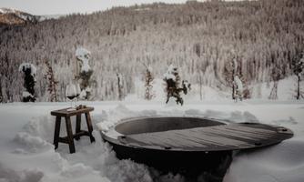A round hot tub is surrounded by snow, with snow-covered trees in the background. Next to the hot tub is a small table with a glass of wine.