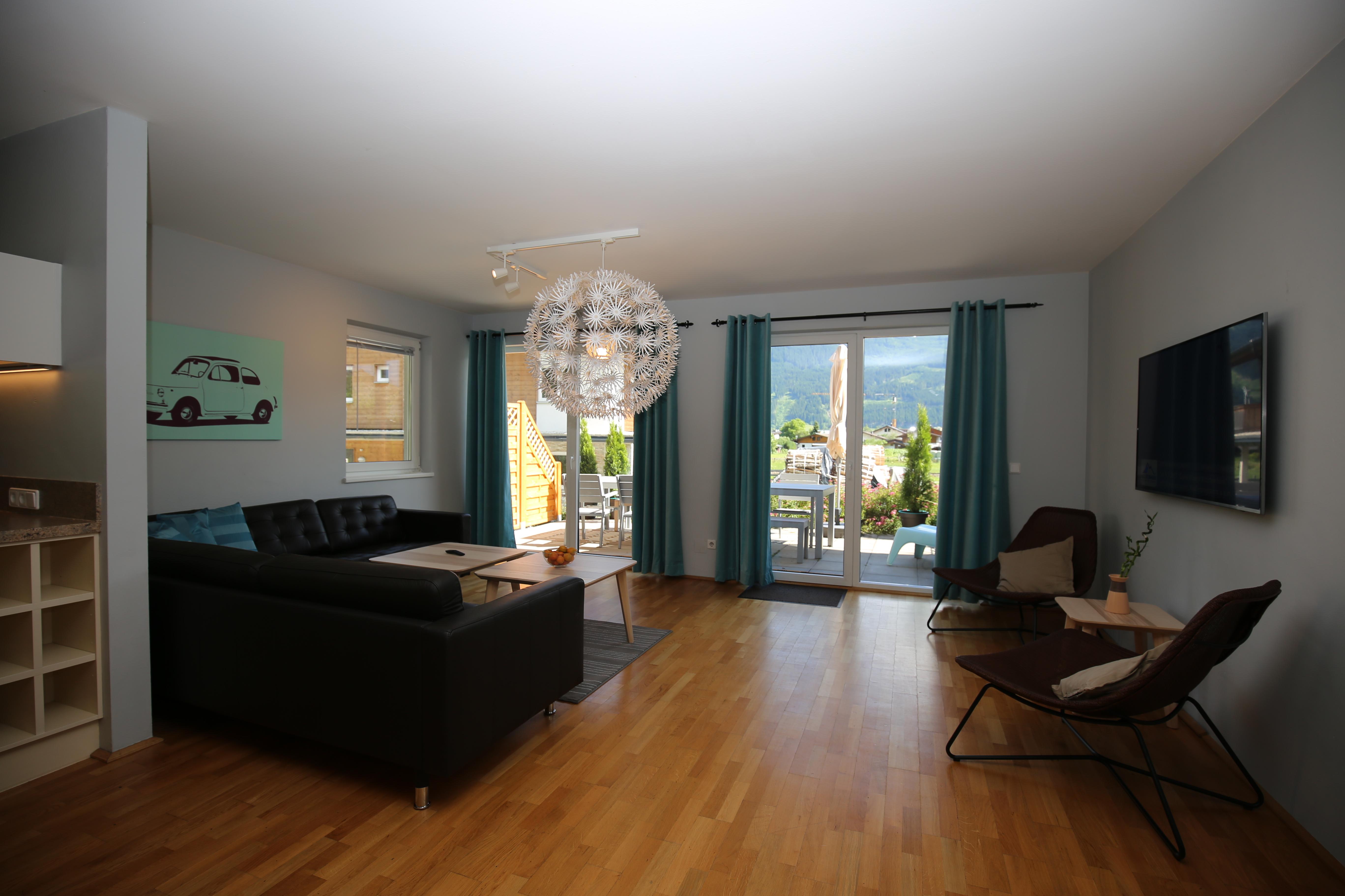 A modern living room with hardwood floors and large windows. It is furnished with a couch, a coffee table, and a television.