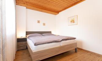 A cozy bedroom with a double bed and wooden ceiling. The walls are light, and there are subtle artworks.