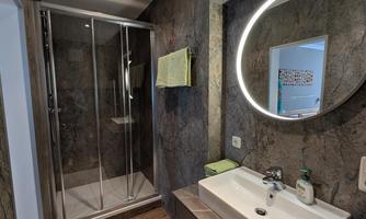 A modern bathroom with a shower and a round mirror. The walls are designed with dark tiles and there are practical shelf spaces.