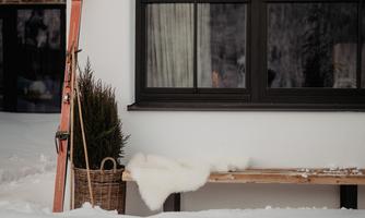 A wintry scene with snow and a cozy bench in front of a house. Skis lean against the wall and a small plant in a basket is visible.
