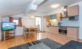 A bright and modern kitchen with a dining area and a television in the living space. The room has wooden flooring and large windows for plenty of daylight.