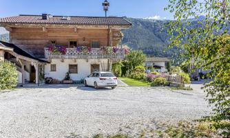 A charming wooden house in a picturesque setting. In the foreground, there is a car on a gravel lot, surrounded by green plants and mountains in the background.