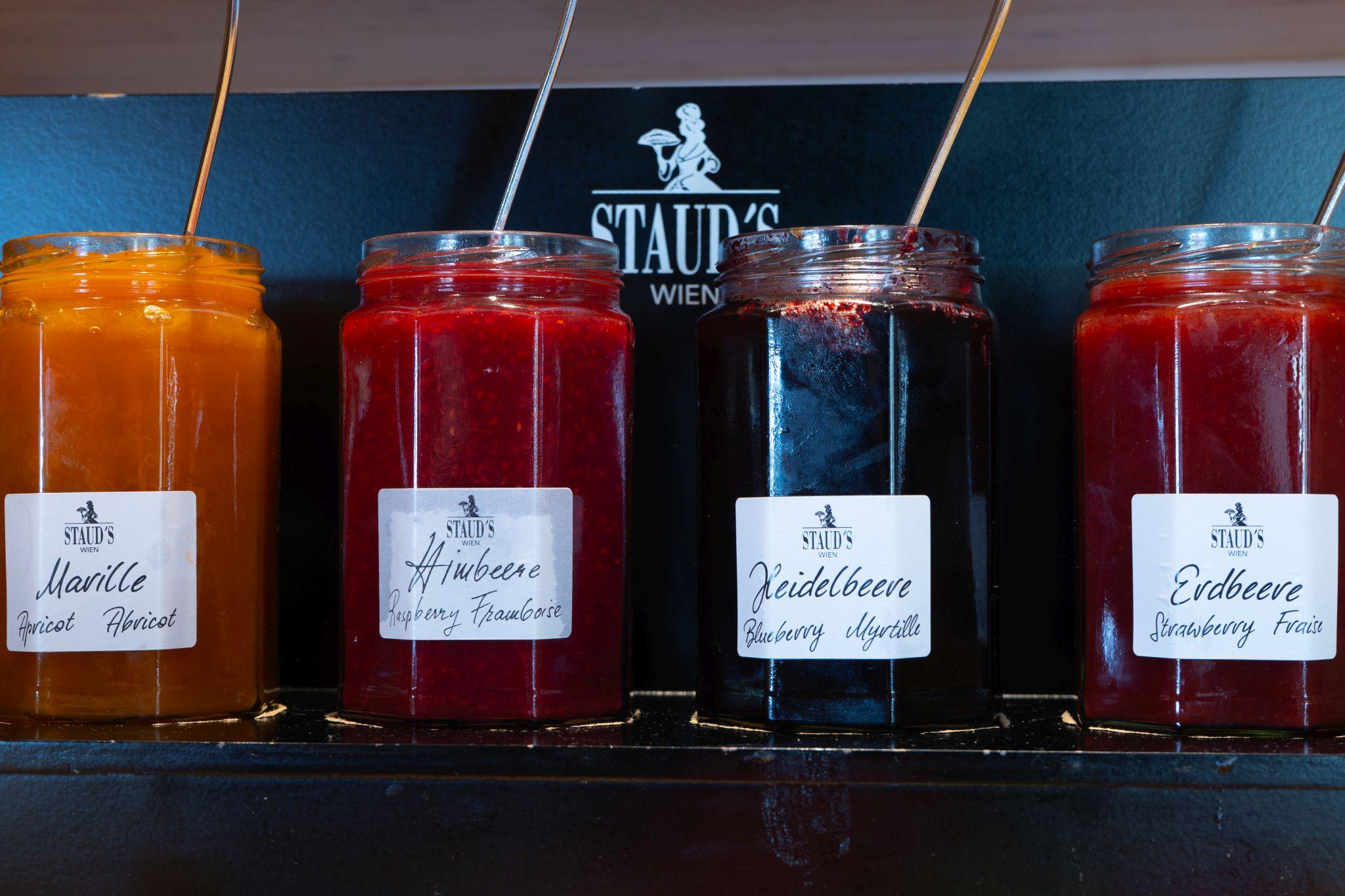 Various jars of jam are standing next to each other, each with a label. The jars contain jam made from apricot, raspberry, blueberry, and strawberry.