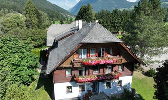 A beautiful house surrounded by trees and mountains. The balcony is adorned with colorful flowers.