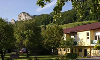 A picturesque building surrounded by trees and green spaces. In the background, impressive, forested mountains can be seen.