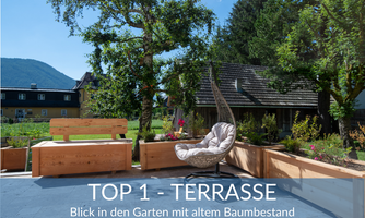 A sunny terrace with modern furniture and a view of a garden with old trees. The surroundings are green and inviting.