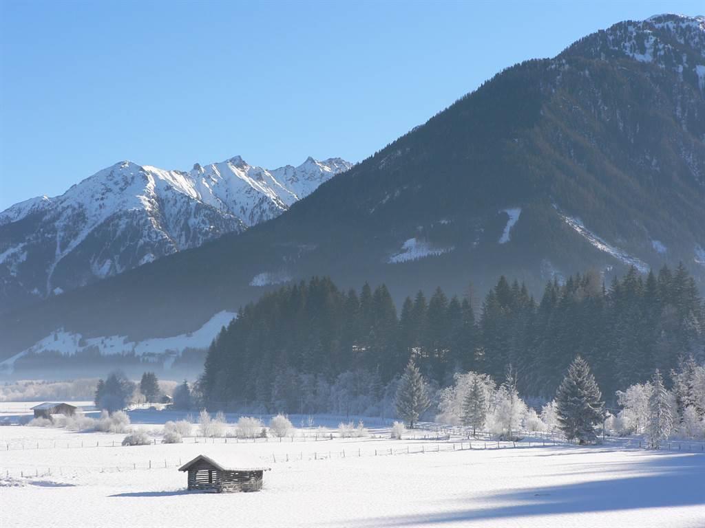 A snowy winter landscape with tall mountains in the background. In the foreground, there is a small black house and some snow-covered trees.