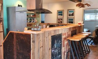 A modern, inviting kitchen with a wooden bar and high stools. Pictures and a board with a menu description hang on the wall.