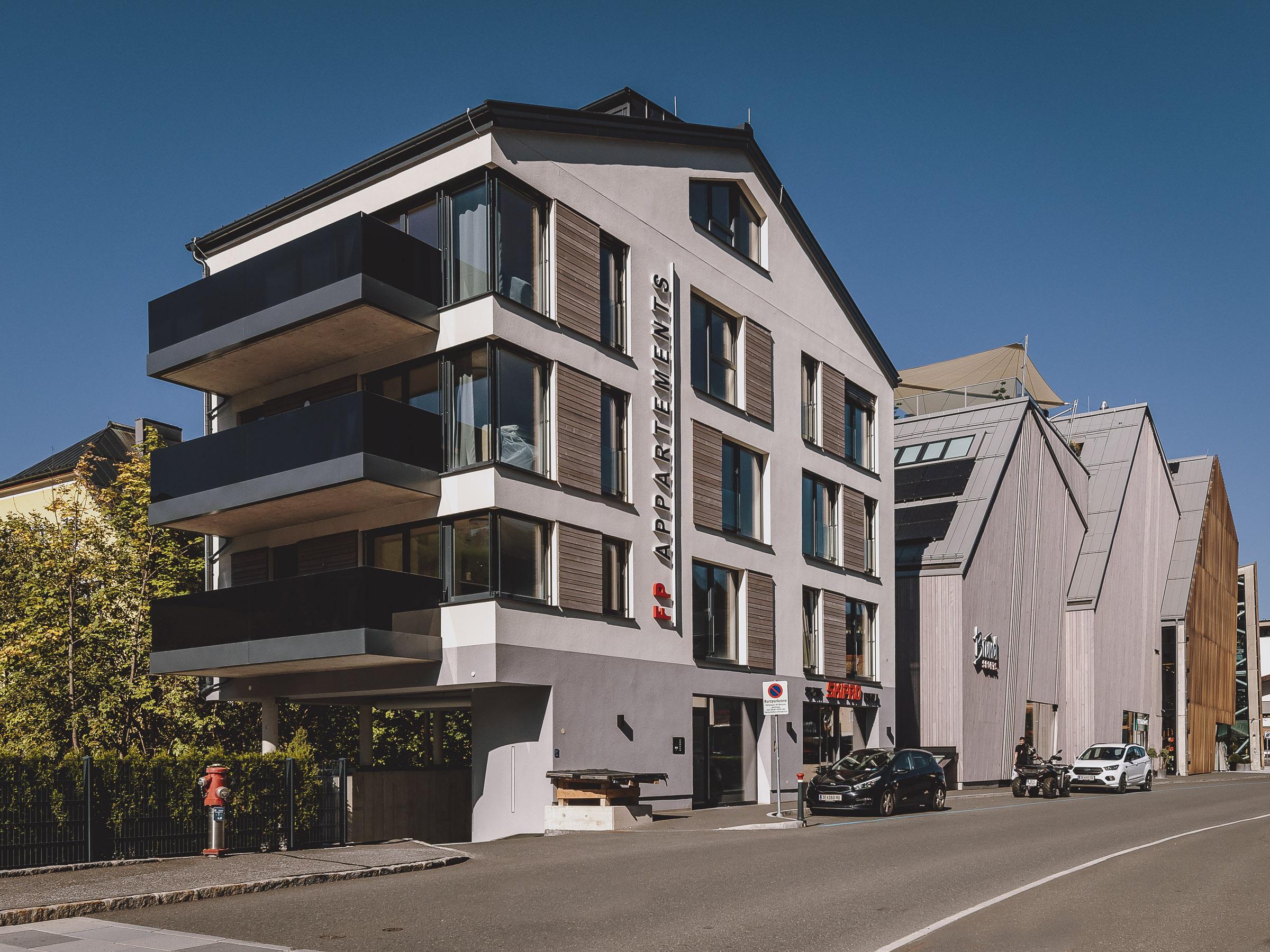A modern building with several balconies and large windows. The street is clearly visible, and some cars are parked.