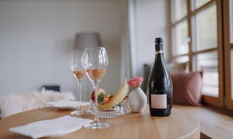 A stylish table with two glasses of sparkling wine, a bottle of sparkling wine, and a small fruit bowl. The room feels bright and inviting with large windows.