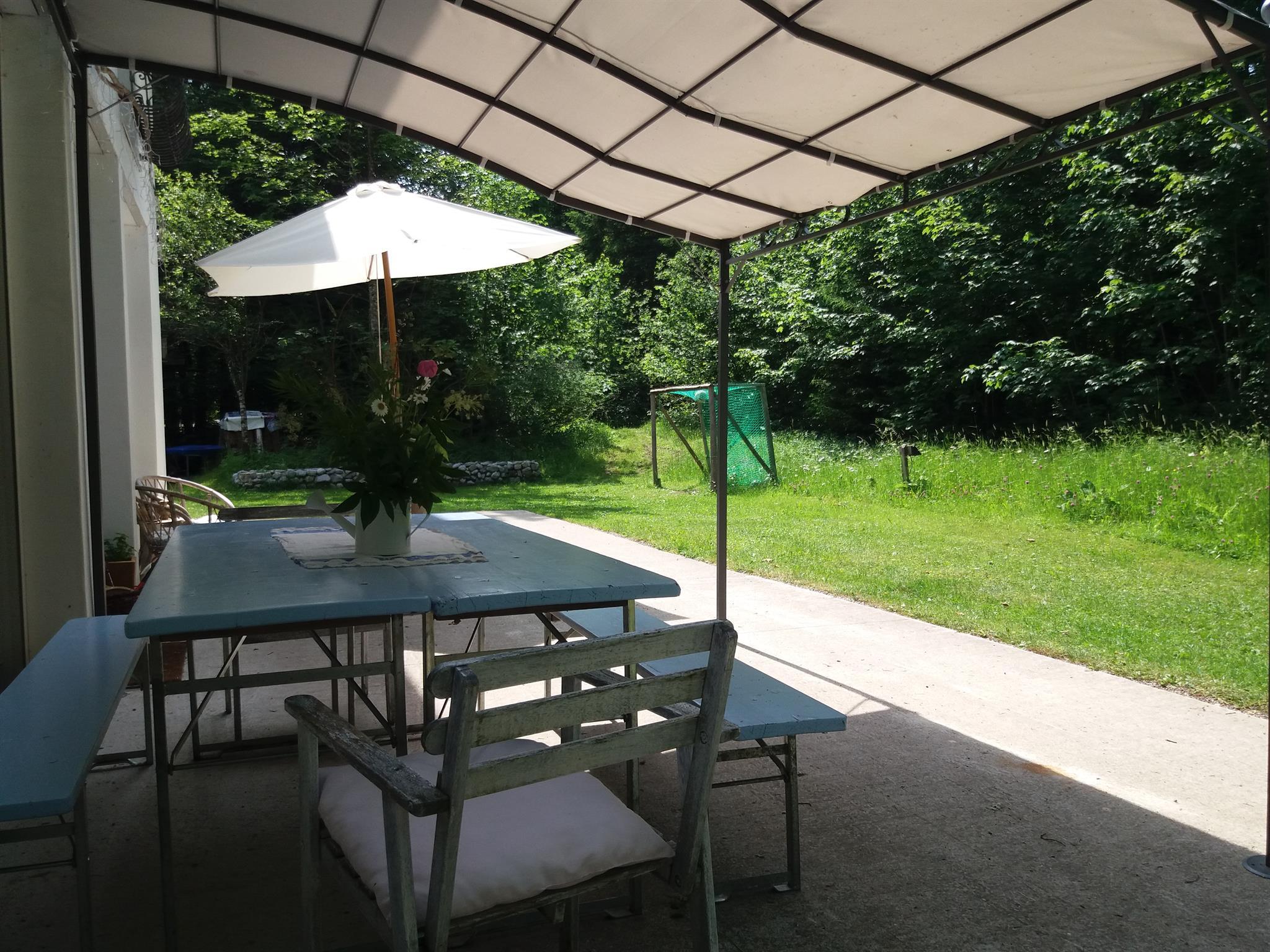 A cozy outdoor area with a table and chairs under a sunshade. In the background, there is a green lawn and trees.
