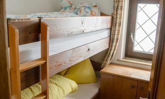 A small room with a bunk bed and a window. The walls are plain and there are some wooden furniture.