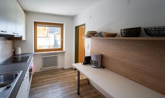 A bright, modern kitchen with a large window and wooden shelves. The cooking area is well-equipped and offers ample space for kitchen utensils.
