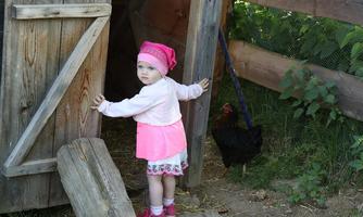 A small girl in a pink outfit is standing by a wooden door. Behind her is a black chicken, and the surroundings are green and rural.