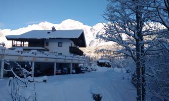 A cozy house in the snow with snow-covered trees and mountains in the background. The sky is clear and blue.