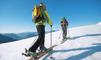 Two skiers are hiking on a snow-covered slope under a clear blue sky. The mountains in the background create an impressive backdrop.
