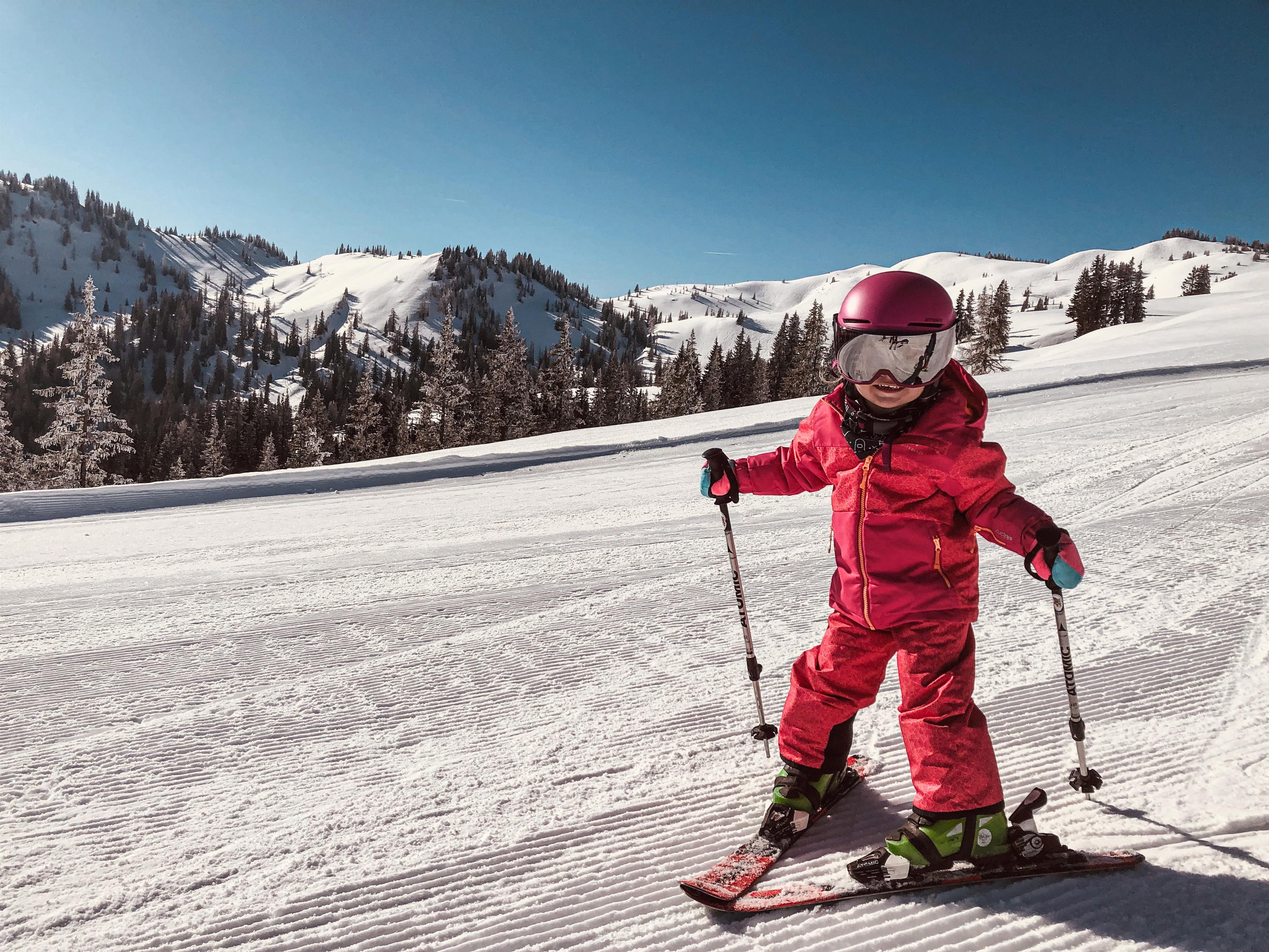 A small child on skis, standing on a snowy slope. In the background, there are snow-covered mountains and a blue sky.