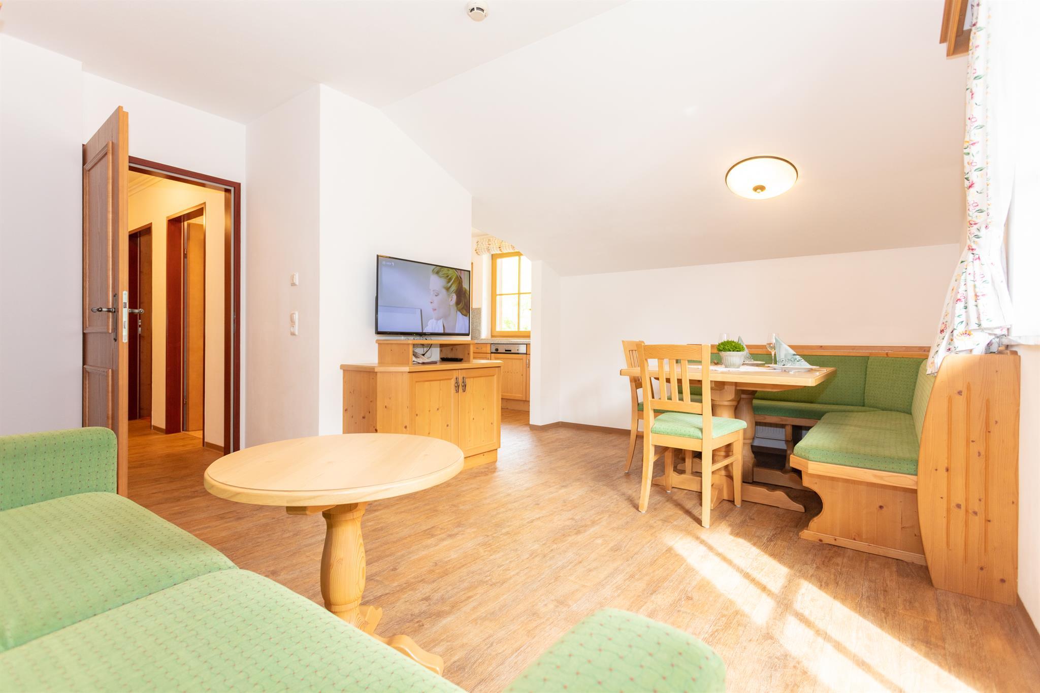 A bright and friendly living room with a cozy green sofa and a table. A dining area with wooden chairs and a TV is also available.