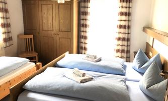 A cozy bedroom with two beds and a bright window front. The furniture is made of wood, and fresh towels are placed on the beds.