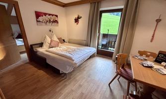A cozy room with a bed, a table, and chairs. Large windows offer a view of the green landscape.
