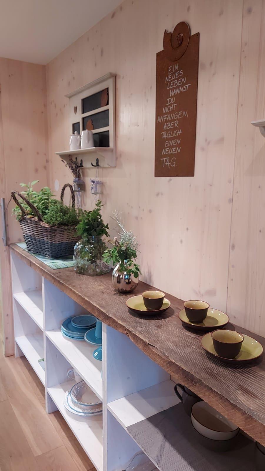A cozy kitchen nook with wooden shelves and a rustic wooden tabletop. On the table, there are some cups and plates next to a decorative plant arrangement.