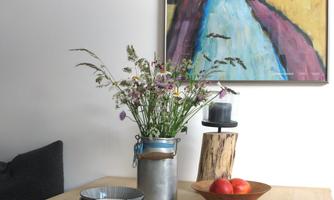 A table with decorative plates and cups, surrounded by fresh flowers in a vase. In the background hangs a colorful painting of a woman.