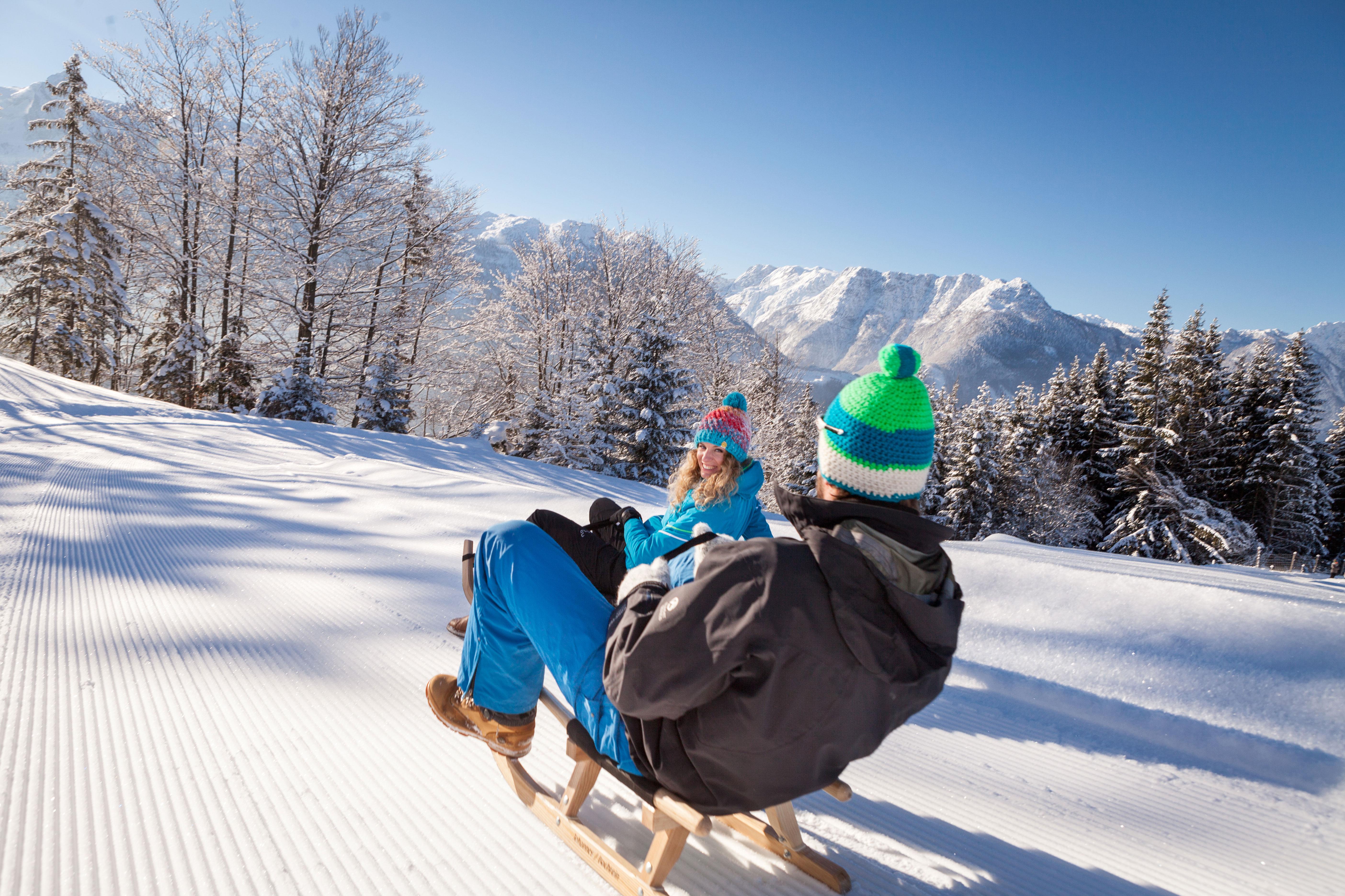 A wintry landscape with snow and high mountains. Two people are sitting on a sled and enjoying the sun.