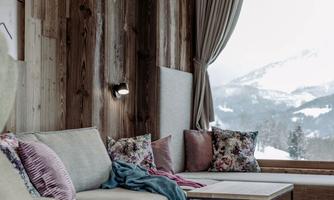 A cozy living room with wooden paneling and large windows. It offers a view of the snow-capped mountains and is stylishly decorated with pillows and blankets.