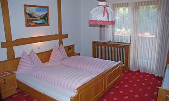 A cozy bedroom with a double bed and two pillows. The walls are made of wood, and the room has a red carpet as well as a large window with curtains.