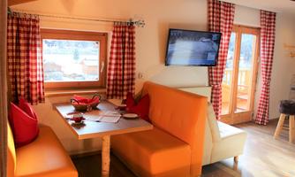 A cozy living kitchen with an orange sofa and wooden table. Large windows provide a view outside and the room is decorated with checkered curtains.