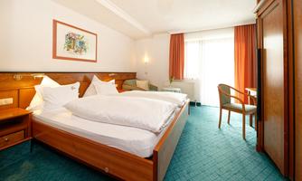 A comfortable hotel room with two single beds and a desk. The room walls are in soft colors, and there are large windows with curtains.