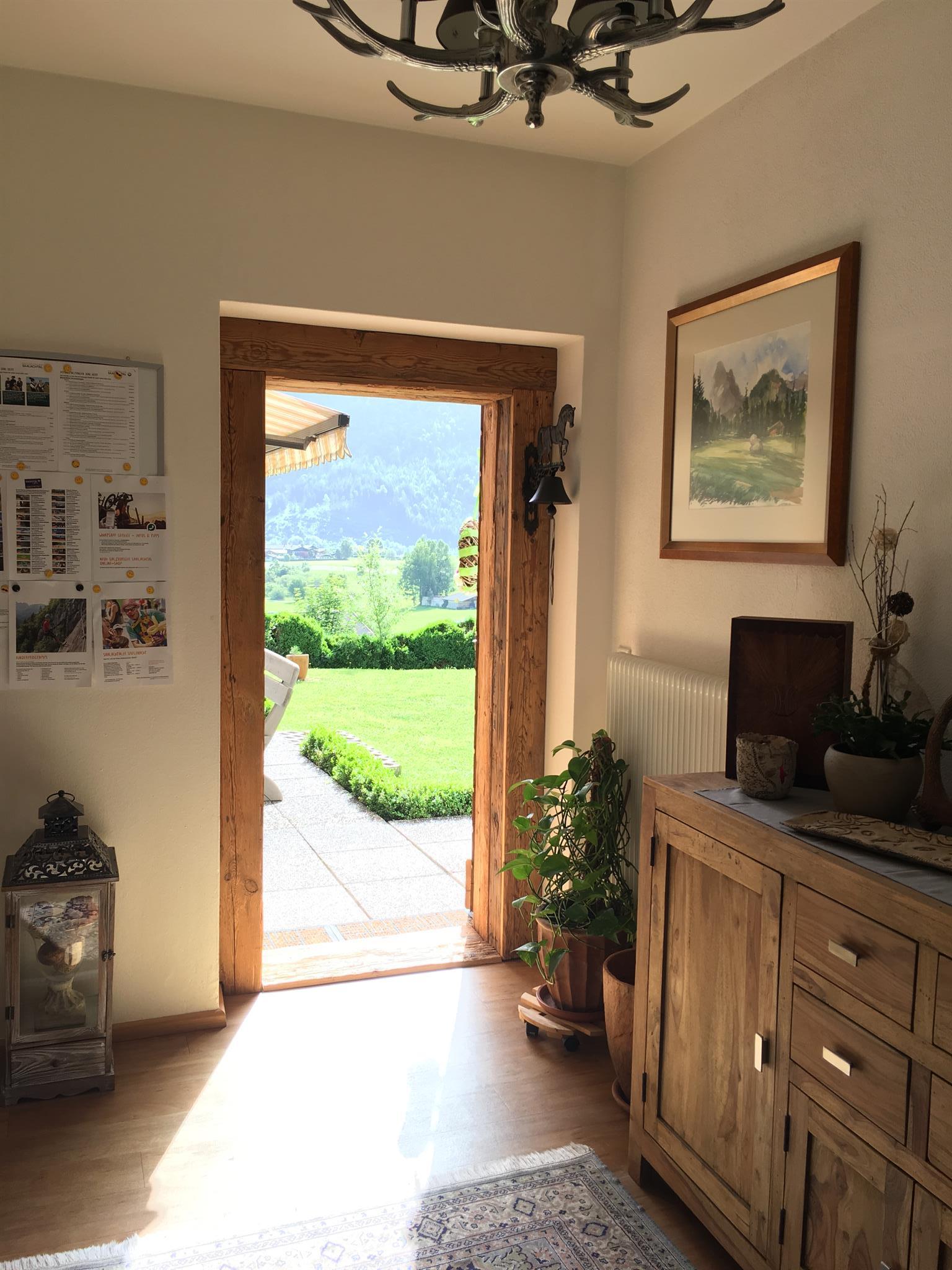 A cozy entrance area with a wooden sideboard and a plant. Through the open door, you can see the garden and the mountains in the background.