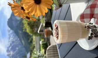 A coffee in a glass sits on a table, surrounded by colorful flowers. In the background, majestic mountains and a clear sky can be seen.