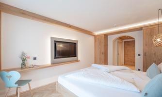 A modern bedroom with a large double bed and elegant wooden décor. There is a television on the wall, and a cozy chair complements the room.