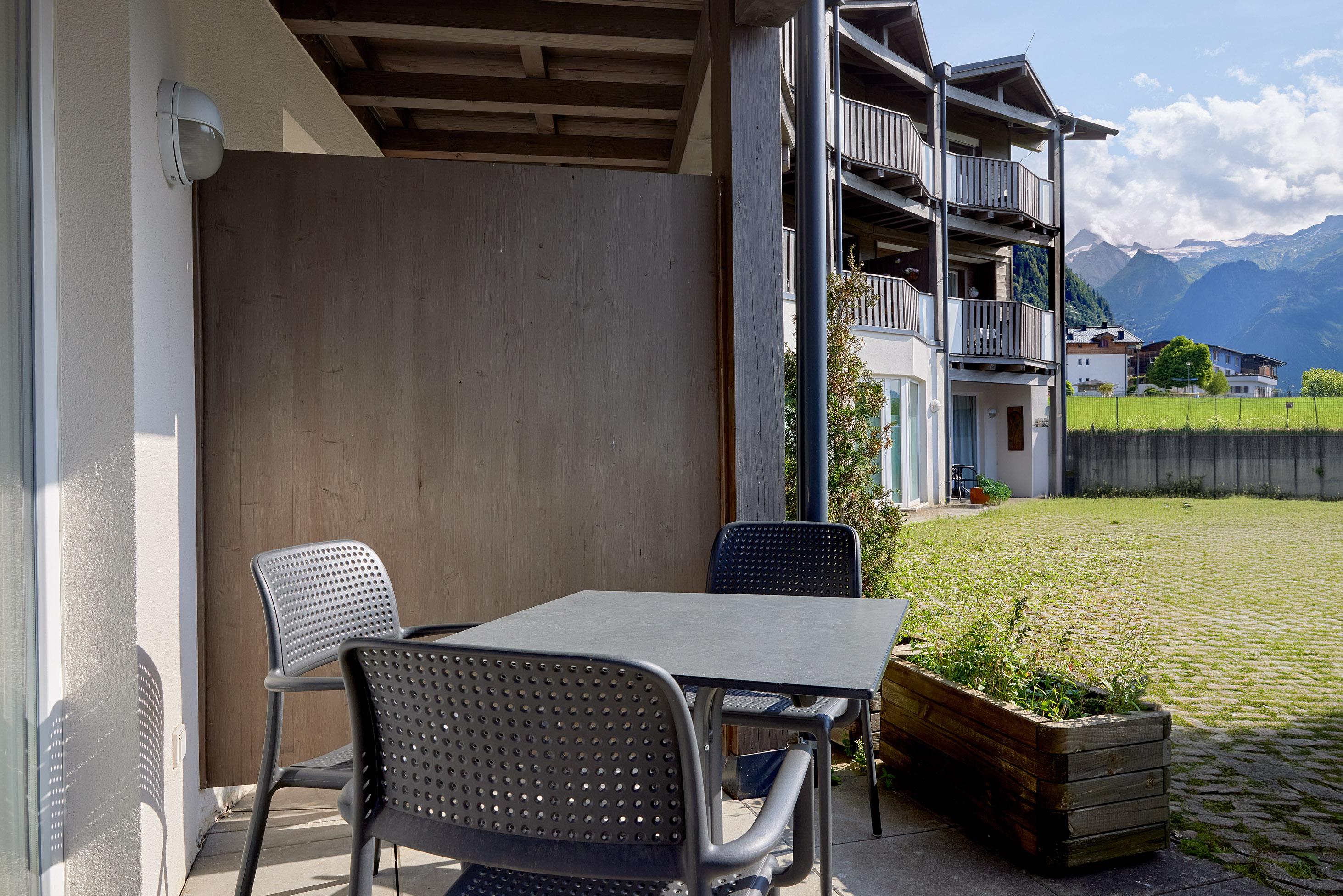 A cozy outdoor area with a table and chairs. In the background, modern buildings and a beautiful landscape with mountains can be seen.