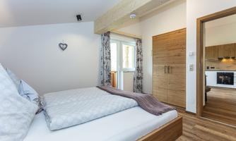 A cozy bedroom with a double bed and wooden details. Daylight falls into the room through a window, which also shows an open door to the kitchen.