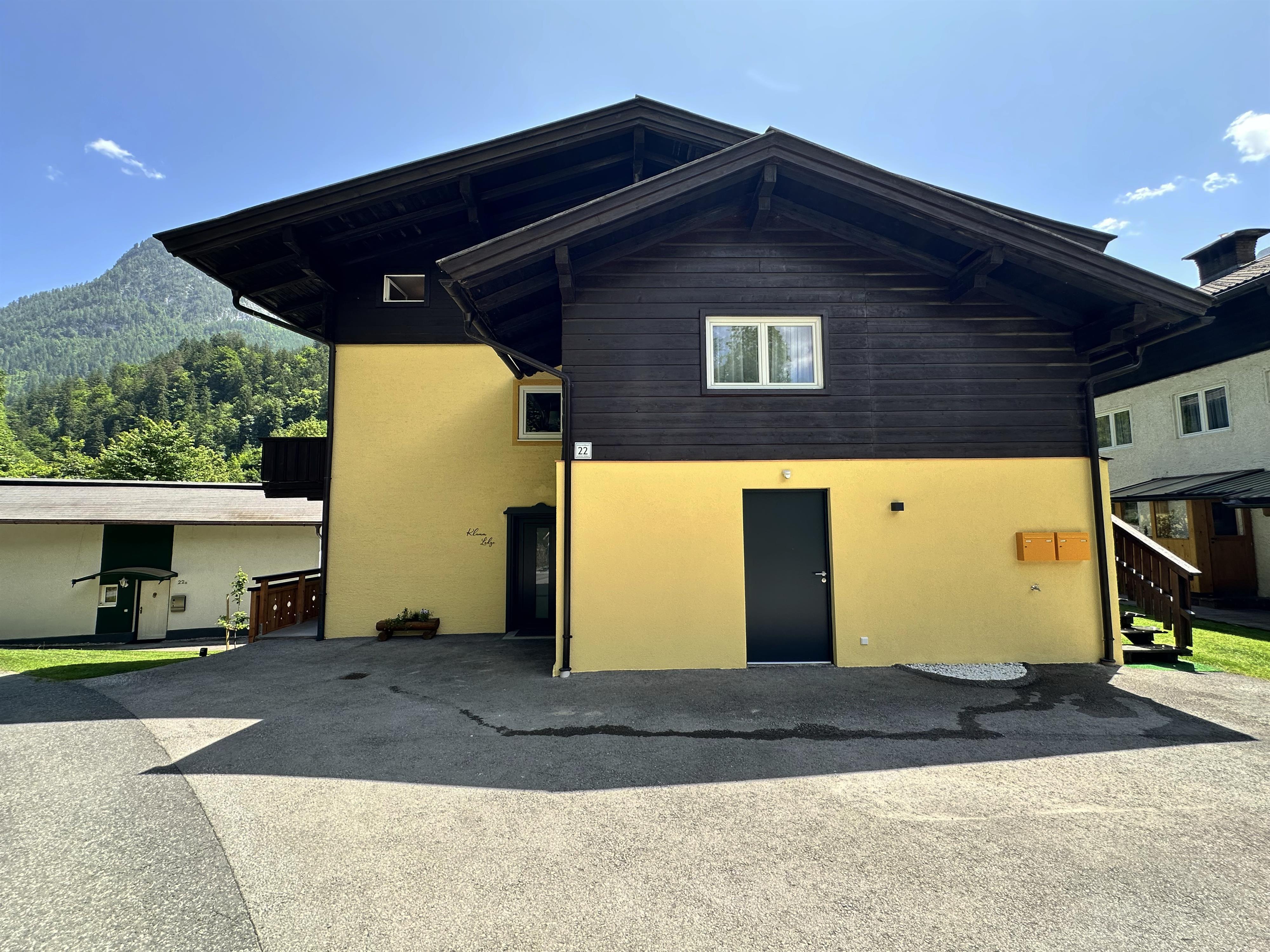 A modern house with a yellow and dark facade in a green environment. In the background, mountains and a blue sky are visible.