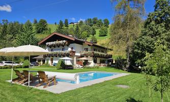 A beautiful house with a balcony, surrounded by trees and meadows. In the foreground, there is a pool with loungers and a sun umbrella.