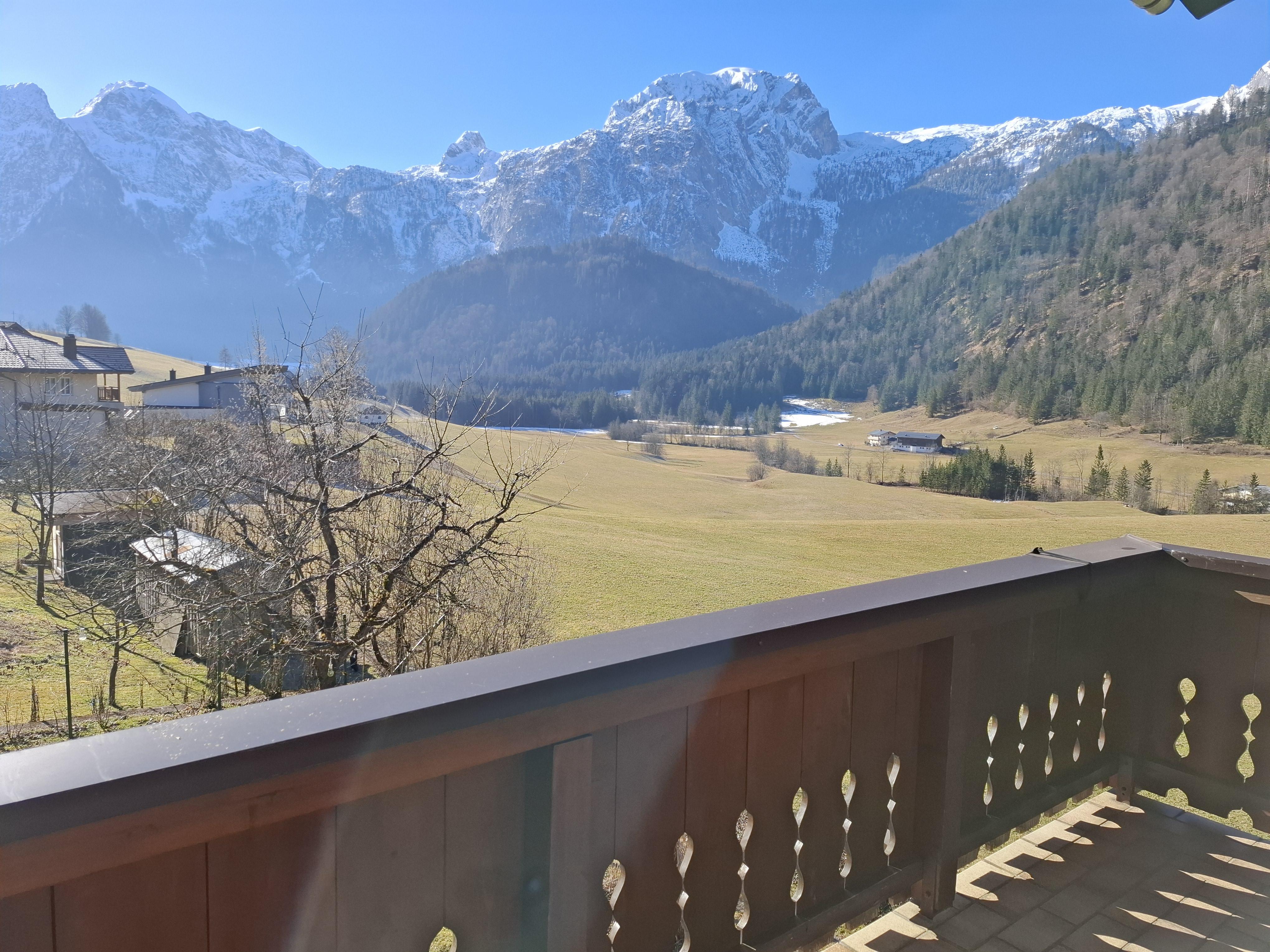 A beautiful view of a vast mountain landscape with snow-covered peaks. In the foreground, there is a balcony with a wooden railing and a green meadow.