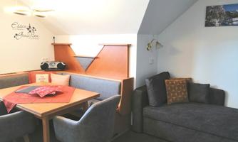 A cozy living area with a dining table, gray chairs, and a comfortable couch. The walls are light, and there is some small decoration.