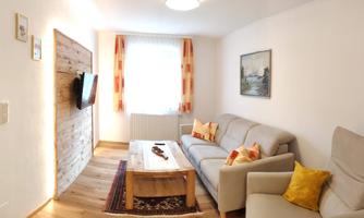 A cozy living room with a bright sofa and a wooden coffee table. The room is decorated with a TV, a painting, and windows with curtains.