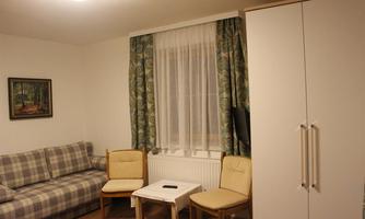 A cozy room with a gray sofa and two chairs. A small table is in the middle, and the room has bright curtains and a wardrobe.