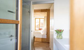 A bright hallway with modern design and wooden accents. In the background, there is a cozy bedroom with a window and a bouquet of flowers on a white table.