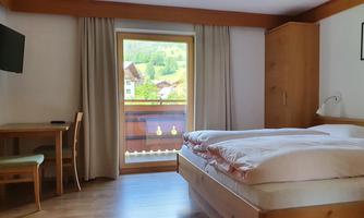 A cozy hotel room with a double bed, a desk, and a television. Through the window, a view of the green landscape is visible.