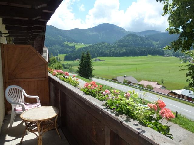 A balcony with a table and chairs, surrounded by colorful flowers. In the background, green meadows and mountains can be seen under a clear sky.