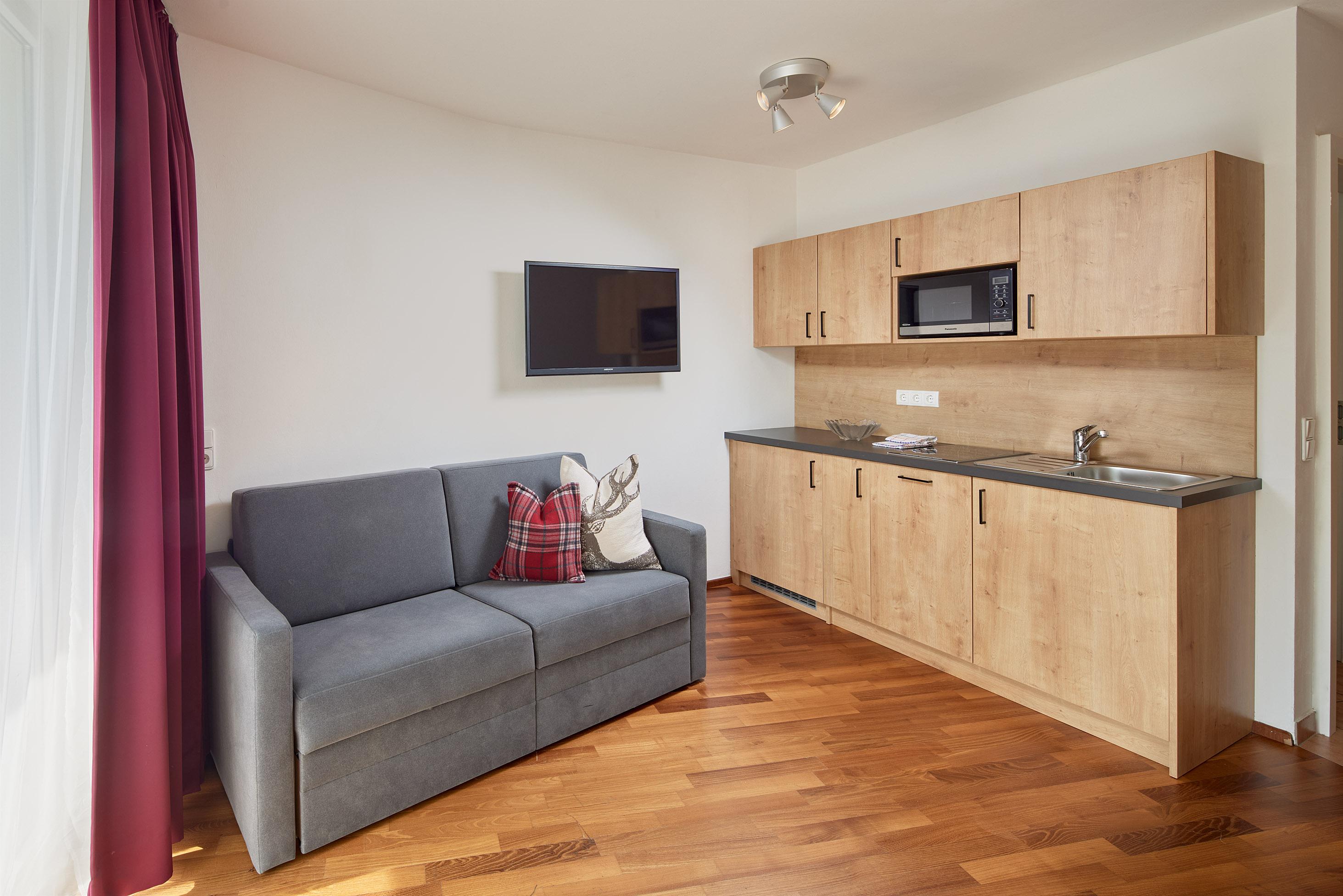 A cozy living room with a gray sofa and a modern kitchenette. The room has wooden flooring and large windows with curtains.