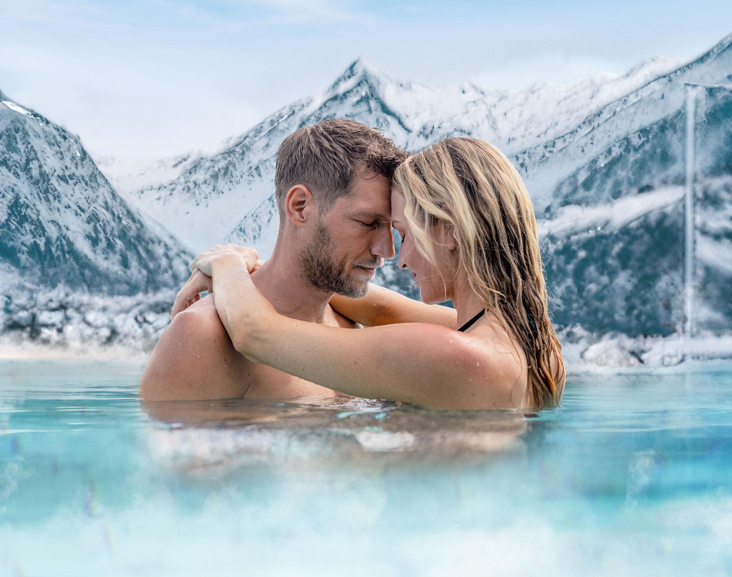 A couple stands in a warm water pool amidst snow-covered mountains. They embrace each other and look lovingly into each other's eyes.