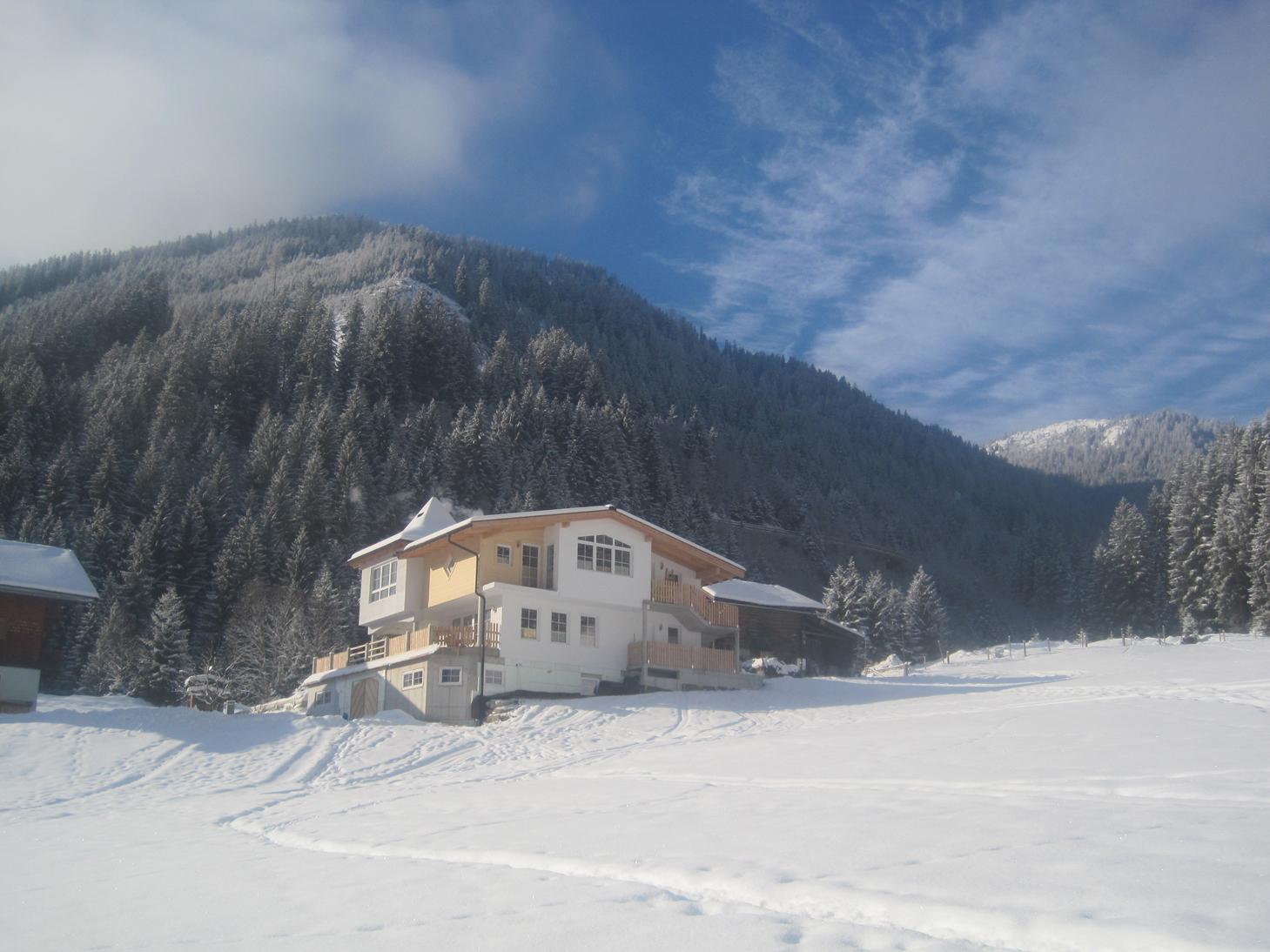 A cozy house in a snowy landscape. In the background, there are wooded mountains and a clear blue sky.